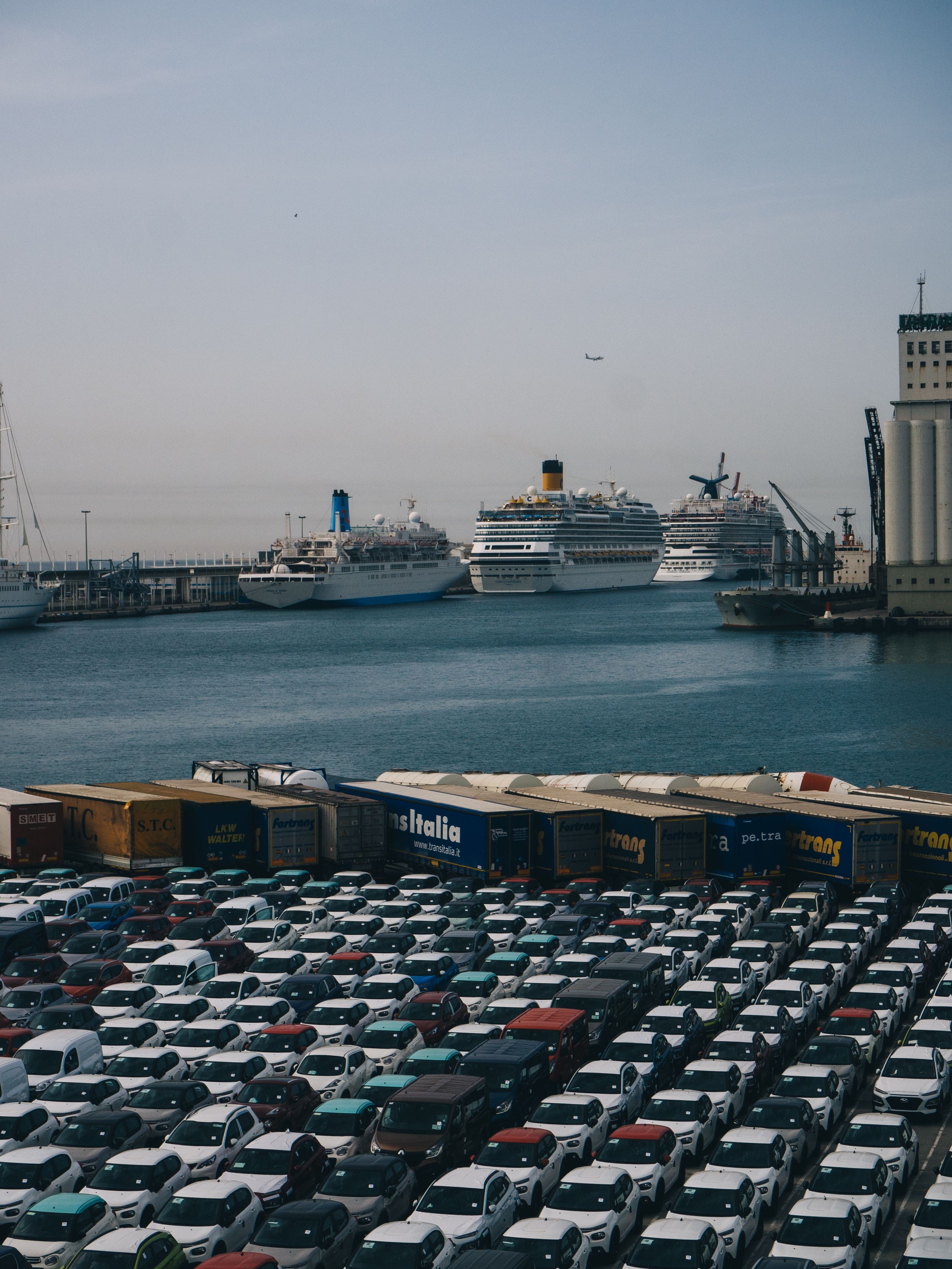 New export cars lined up at a port with cruise ships and freight trailers in the background